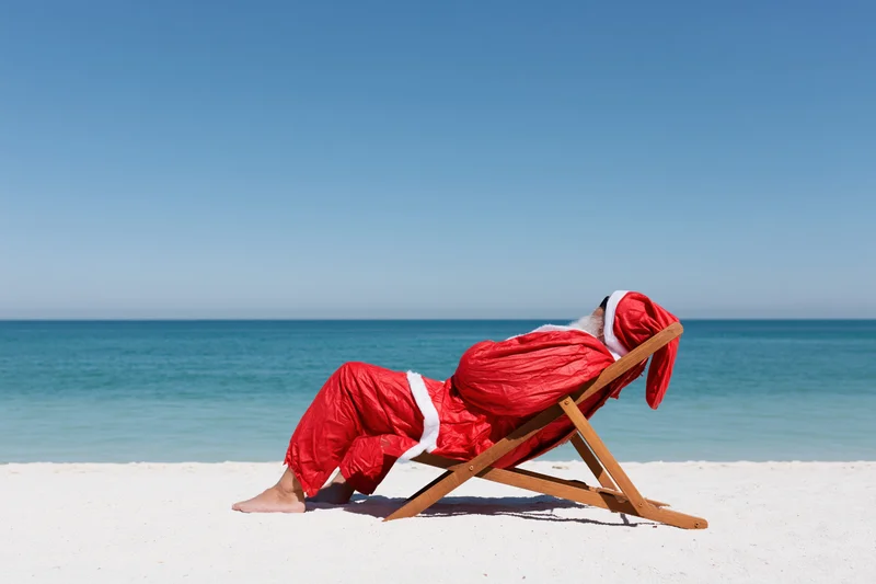 Santa reclining in a deckchair on a sandy beach beside the ocean