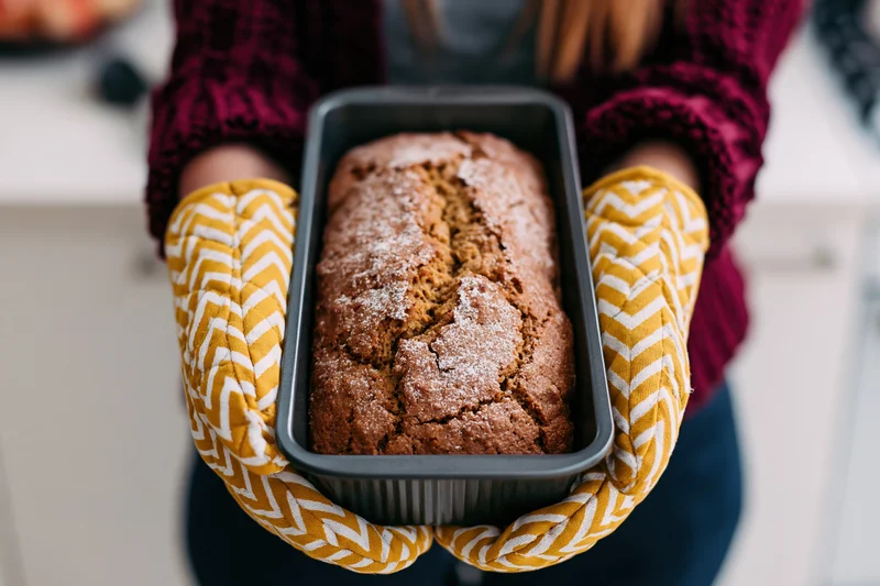 Woman holding freshly baked loaf in baking pan with yellow patterned oven mitts in kitchen