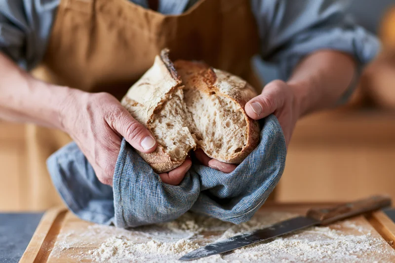 Baker holding freshly baked sourdough bread with hands wrapped in cloth over a floured cutting board