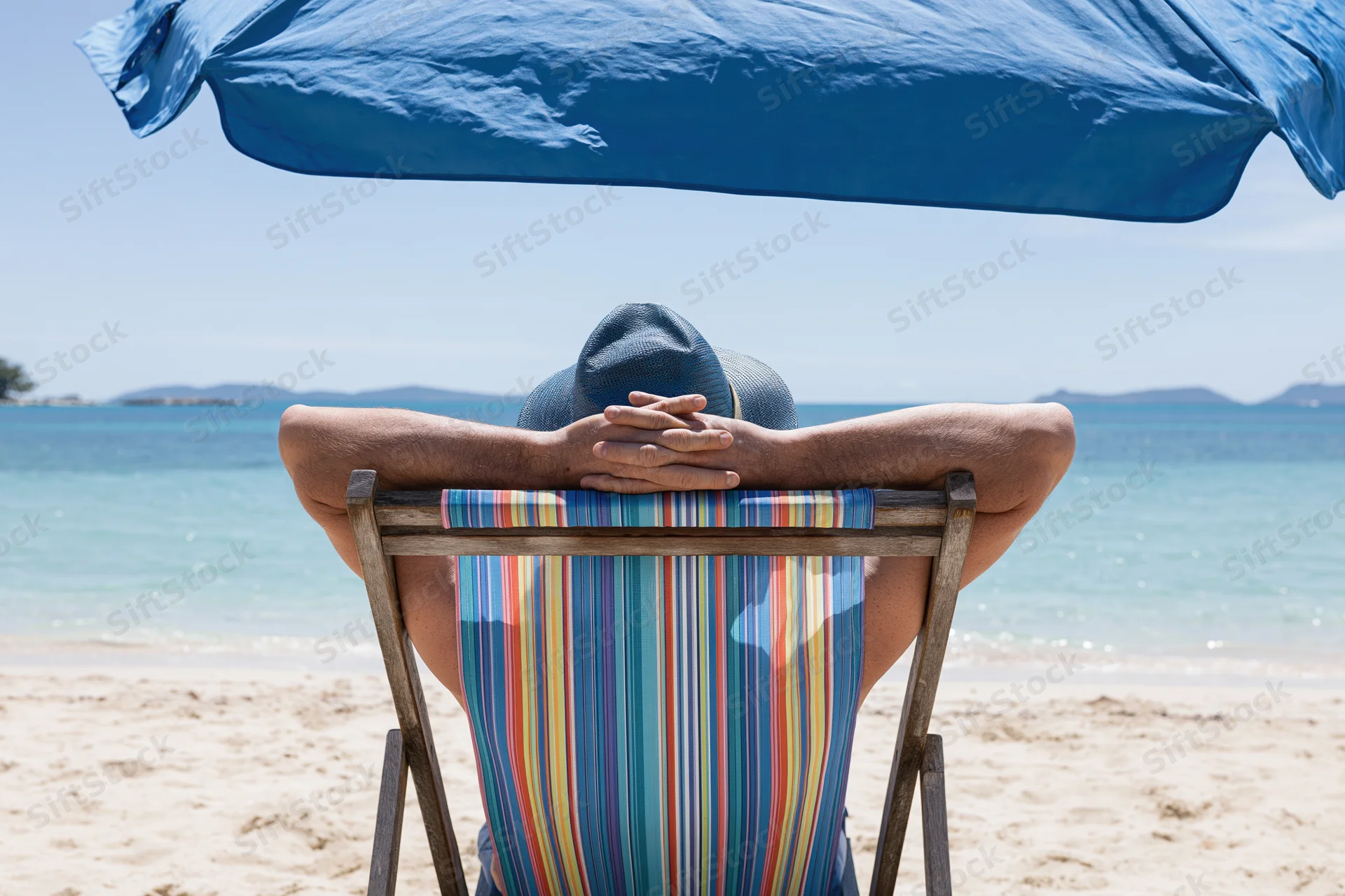 Man reclining in striped deckchair under blue umbrella on sandy beach facing ocean