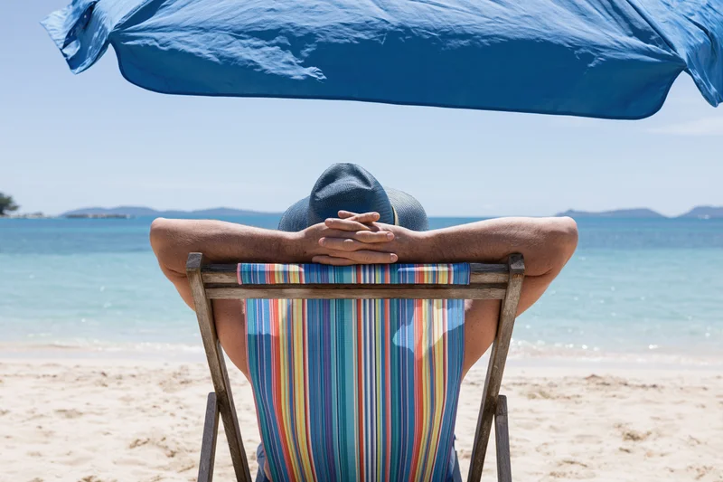 Man reclining in striped deckchair under blue umbrella on sandy beach facing ocean