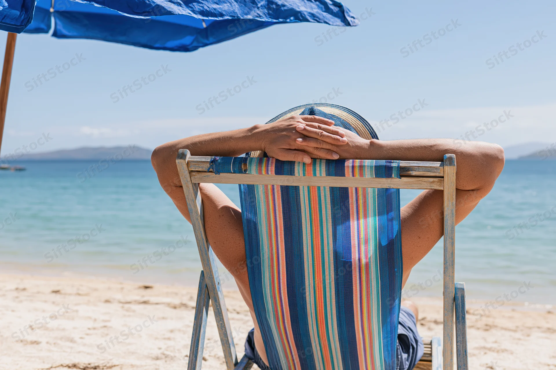 Woman reclining in striped deckchair under blue umbrella on sandy beach by ocean