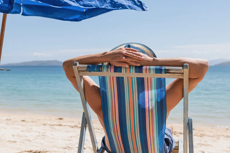 Woman reclining in striped deckchair under blue umbrella on sandy beach by ocean