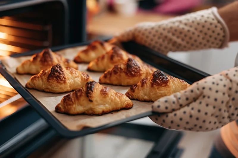 Hands in polka-dot oven mitts pulling a tray of golden baked croissants from an oven