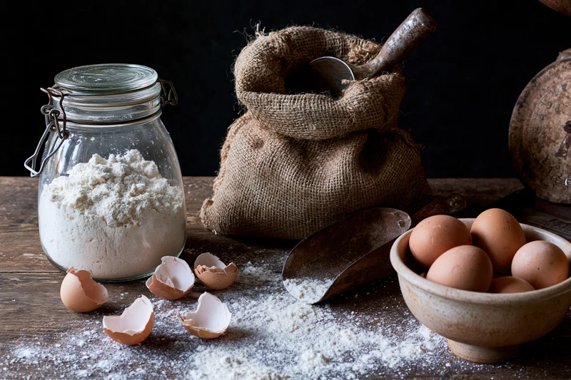 Baking ingredients arranged on a wooden table with flour, eggs, and a burlap sack