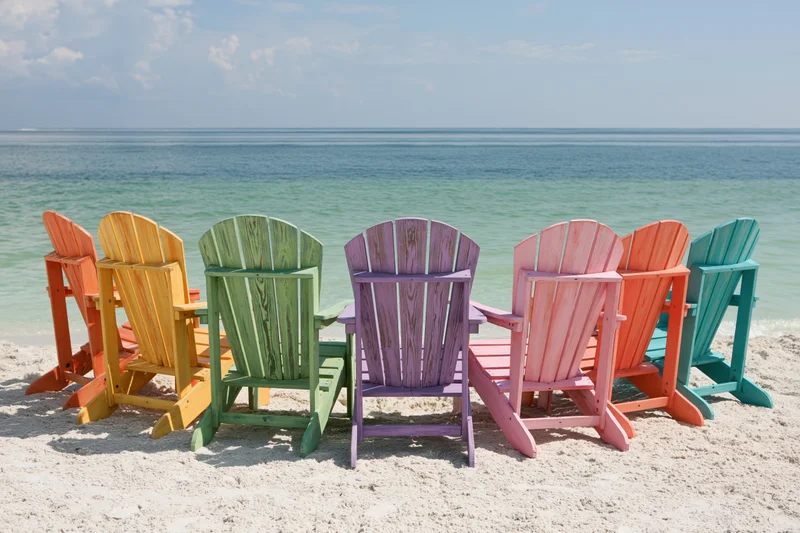 Colorful adirondack chairs arranged on sandy beach facing ocean under sunny sky