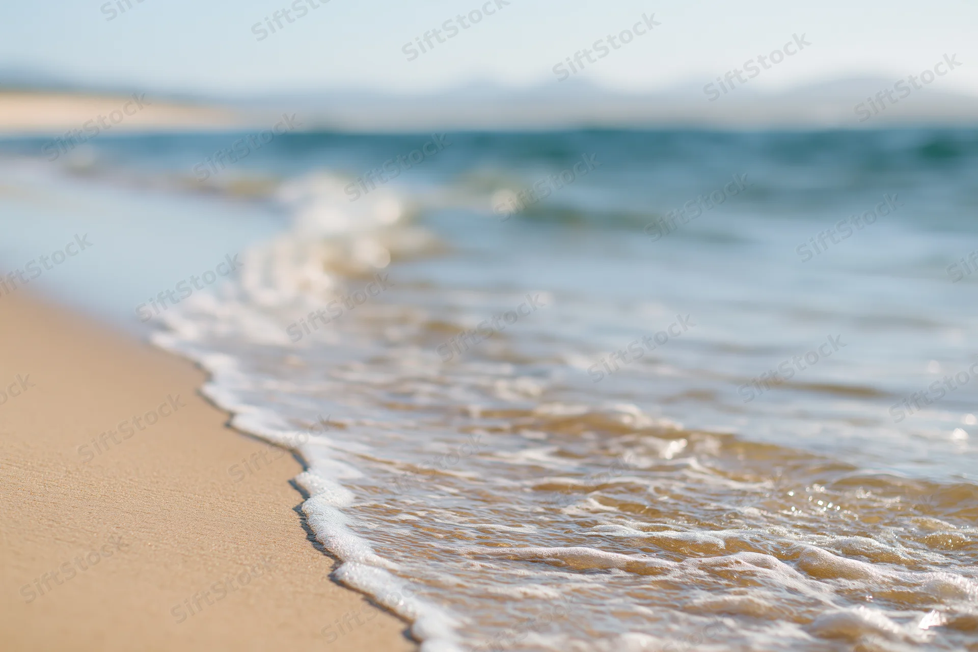 Gentle wave washing over sandy beach at shoreline with distant mountains and clear sky