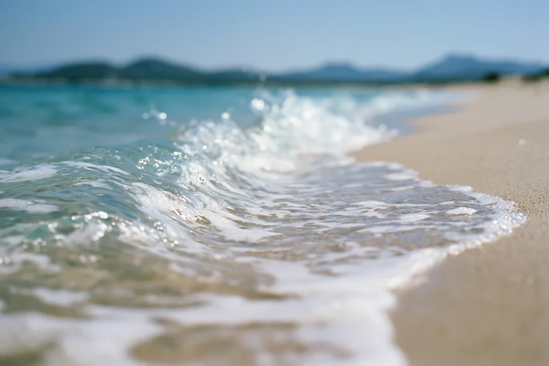 Gentle wave washing onto sandy beach with distant mountains under clear sky