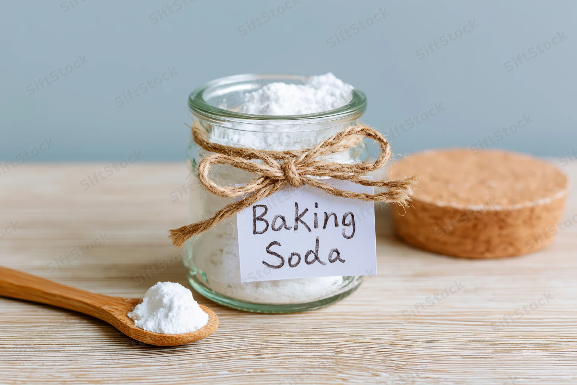 Glass jar filled with white powder labeled baking soda on wooden surface with wooden spoon and cork lid beside it