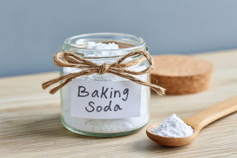 Glass jar filled with white powder labeled baking soda on wooden surface with wooden spoon beside it
