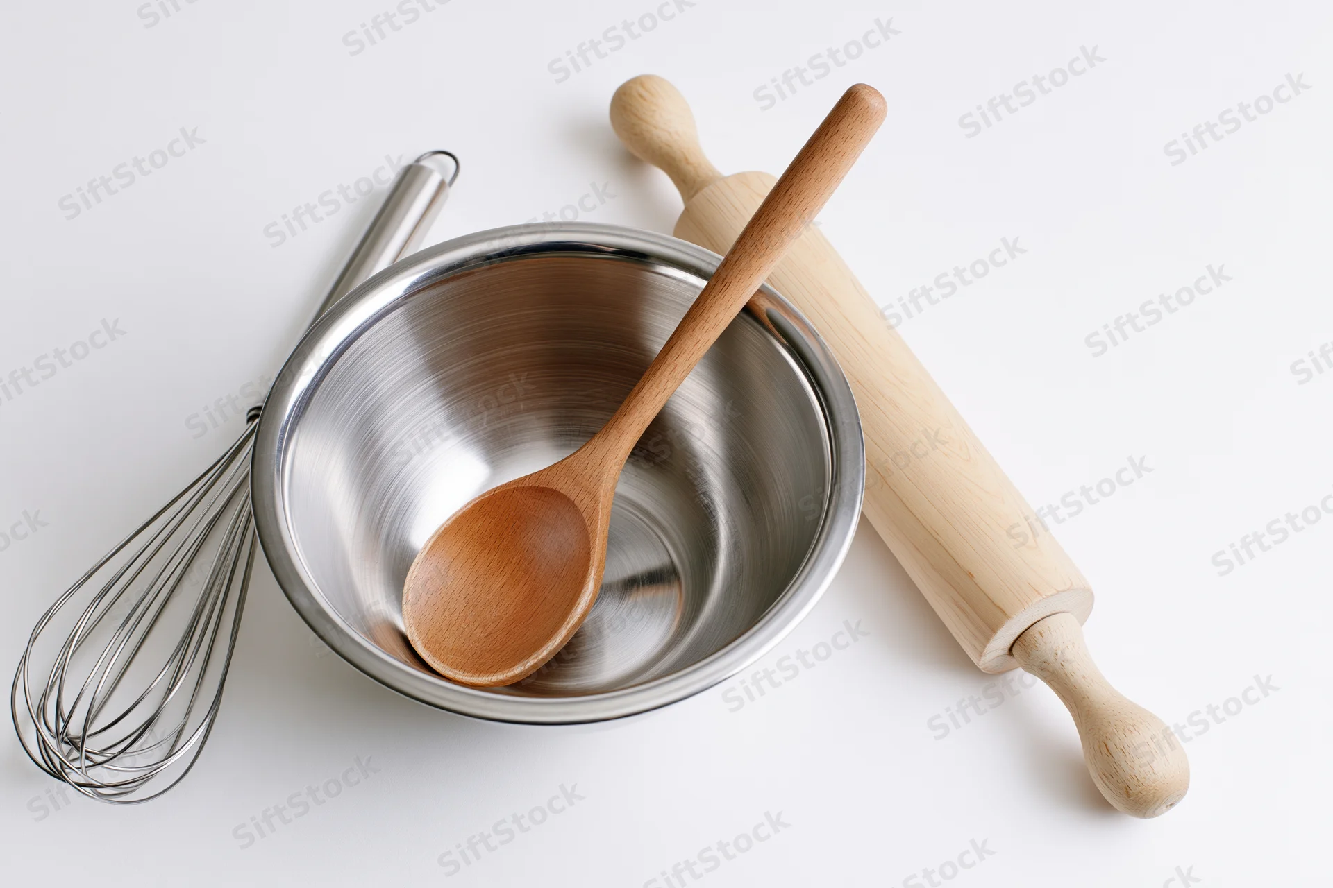 Stainless steel bowl with wooden spoon, whisk and rolling pin arranged on white surface