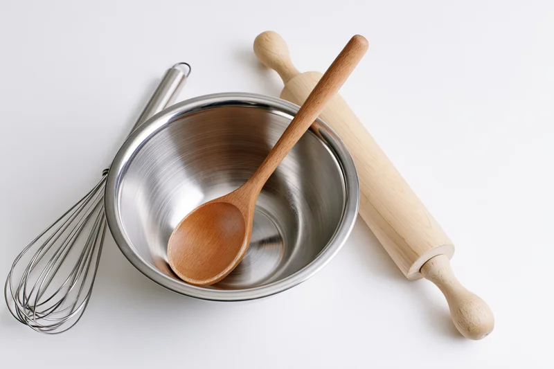 Stainless steel bowl with wooden spoon, whisk and rolling pin arranged on white surface