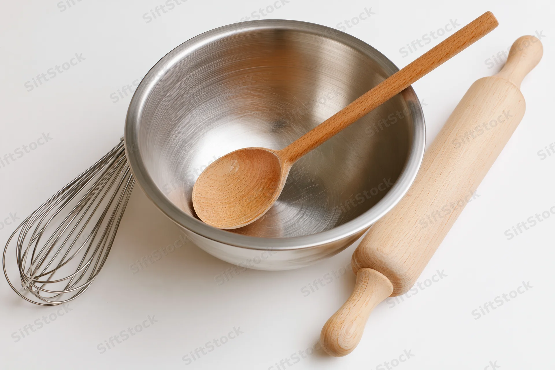 Stainless steel mixing bowl with wooden spoon, whisk and rolling pin on white surface