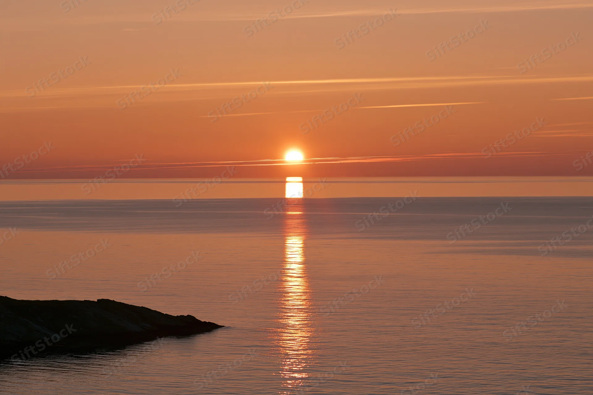Sunset over calm ocean with sun reflecting on water near rocky shore