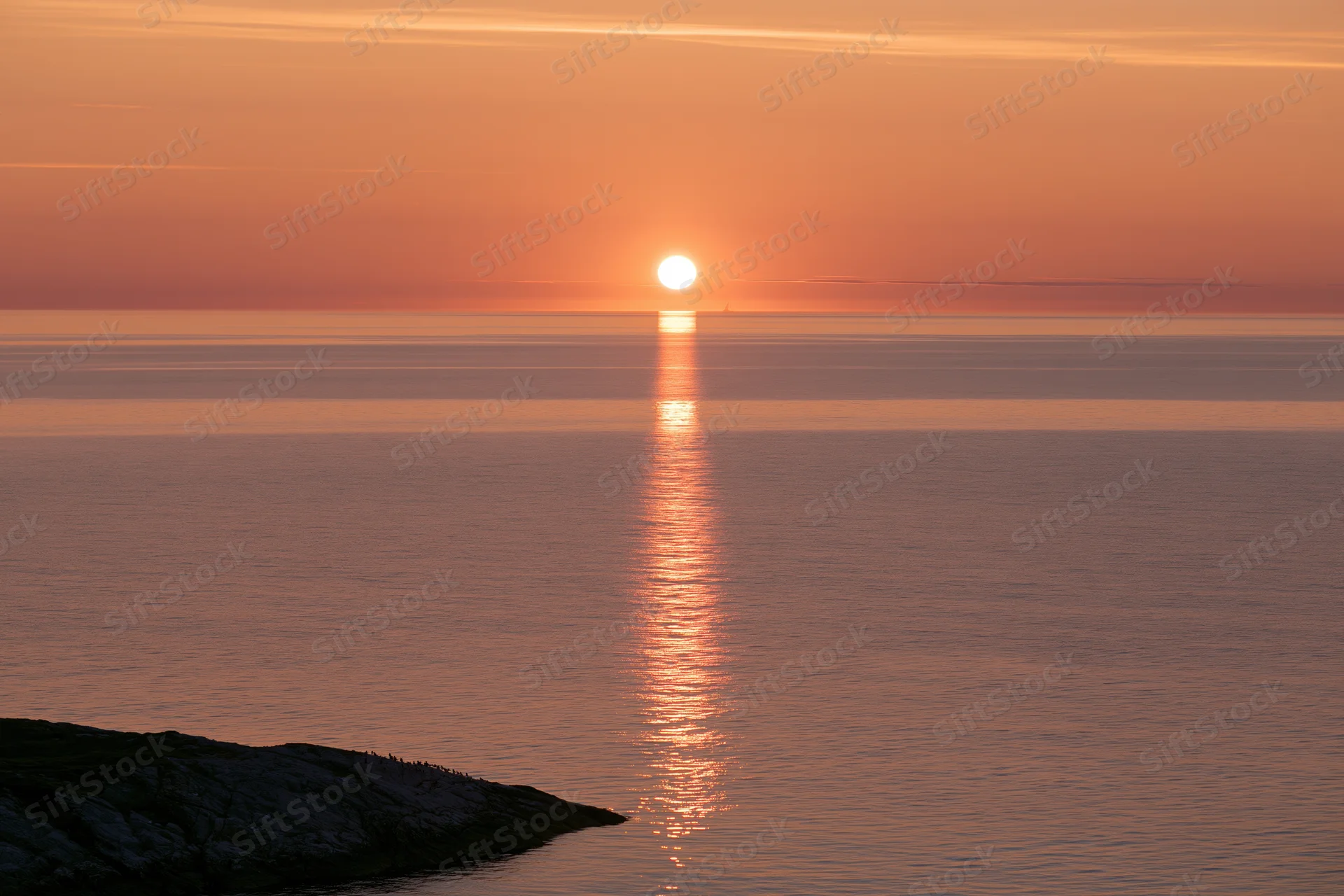 Sunset over calm ocean with sun reflecting on water near rocky shore