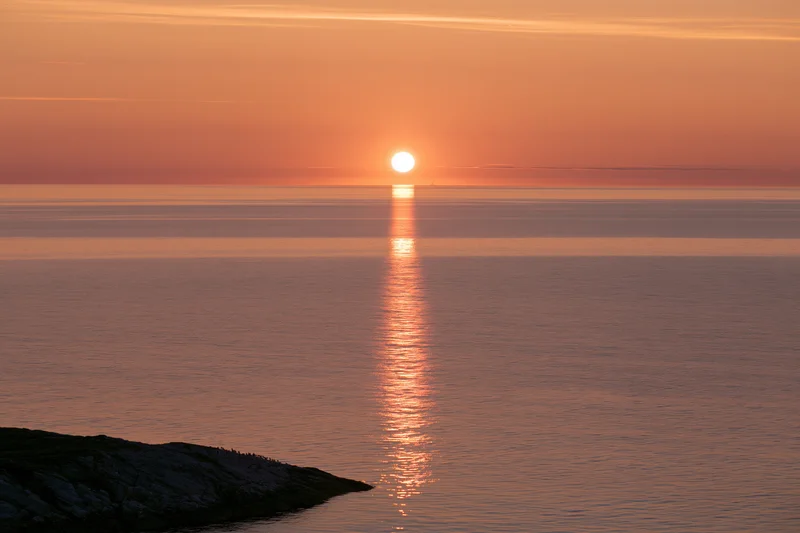 Sunset over calm ocean with sun reflecting on water near rocky shore