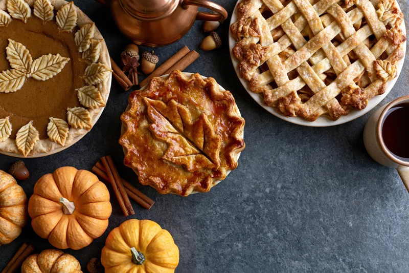 Three pumpkin pies with decorative crusts and autumnal garnishes arranged on a dark table with pumpkins, cinnamon sticks, and a copper kettle