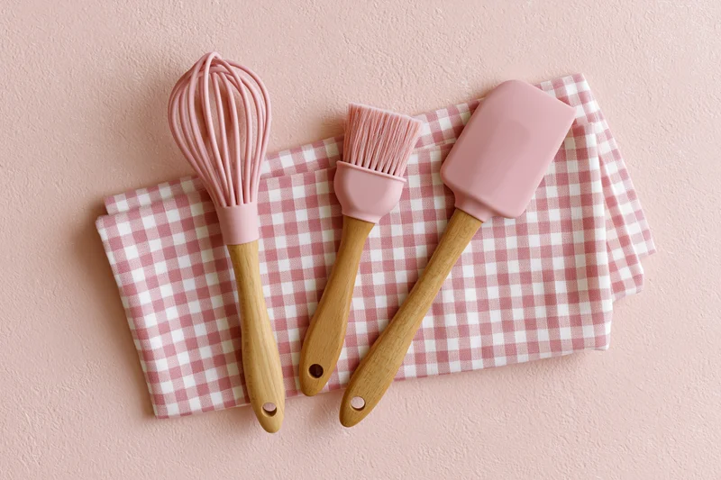 Pink silicone kitchen utensils with wooden handles arranged on a checkered napkin on a pastel pink surface