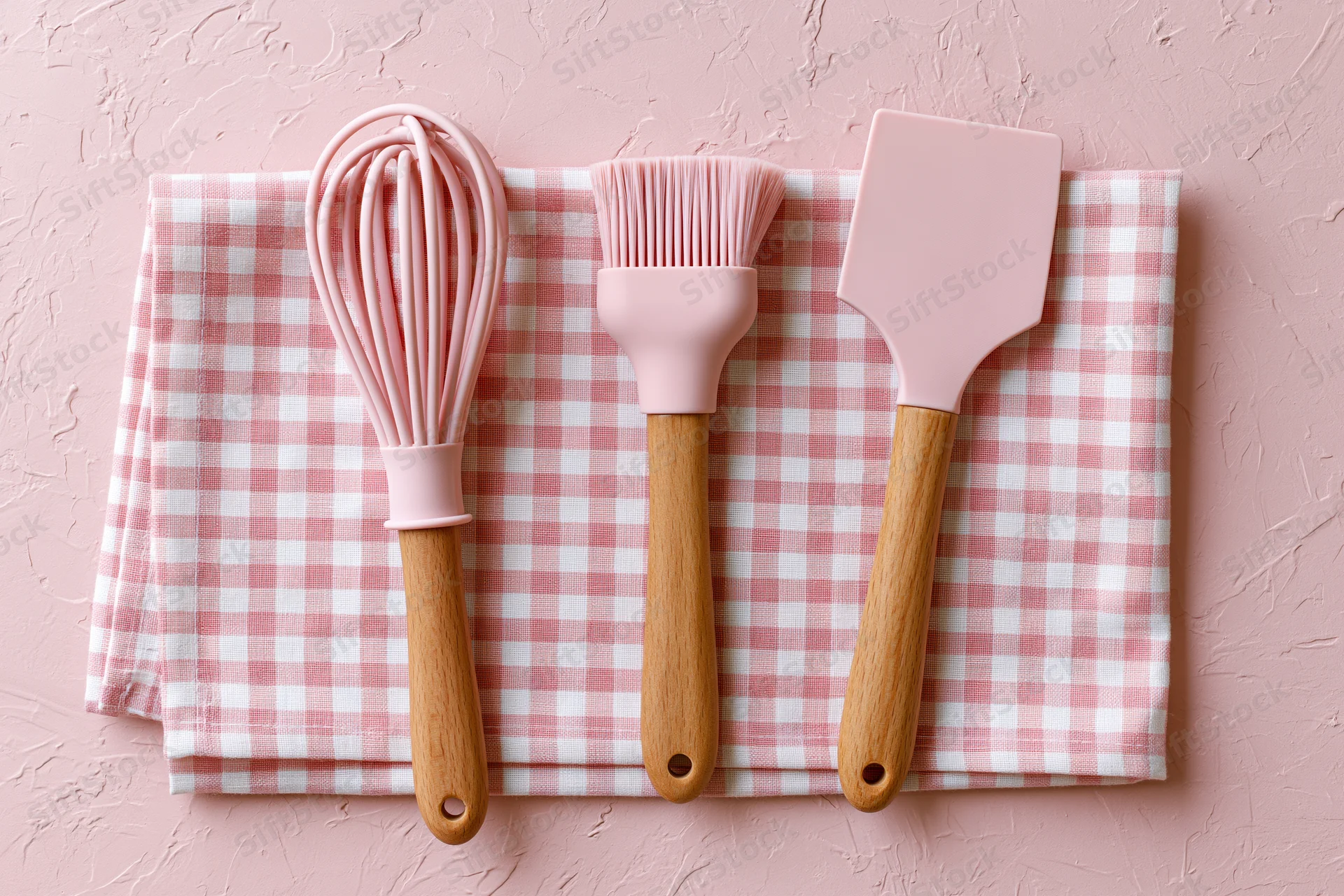 Pink silicone and wooden kitchen utensils arranged on a checkered cloth over a textured pink surface