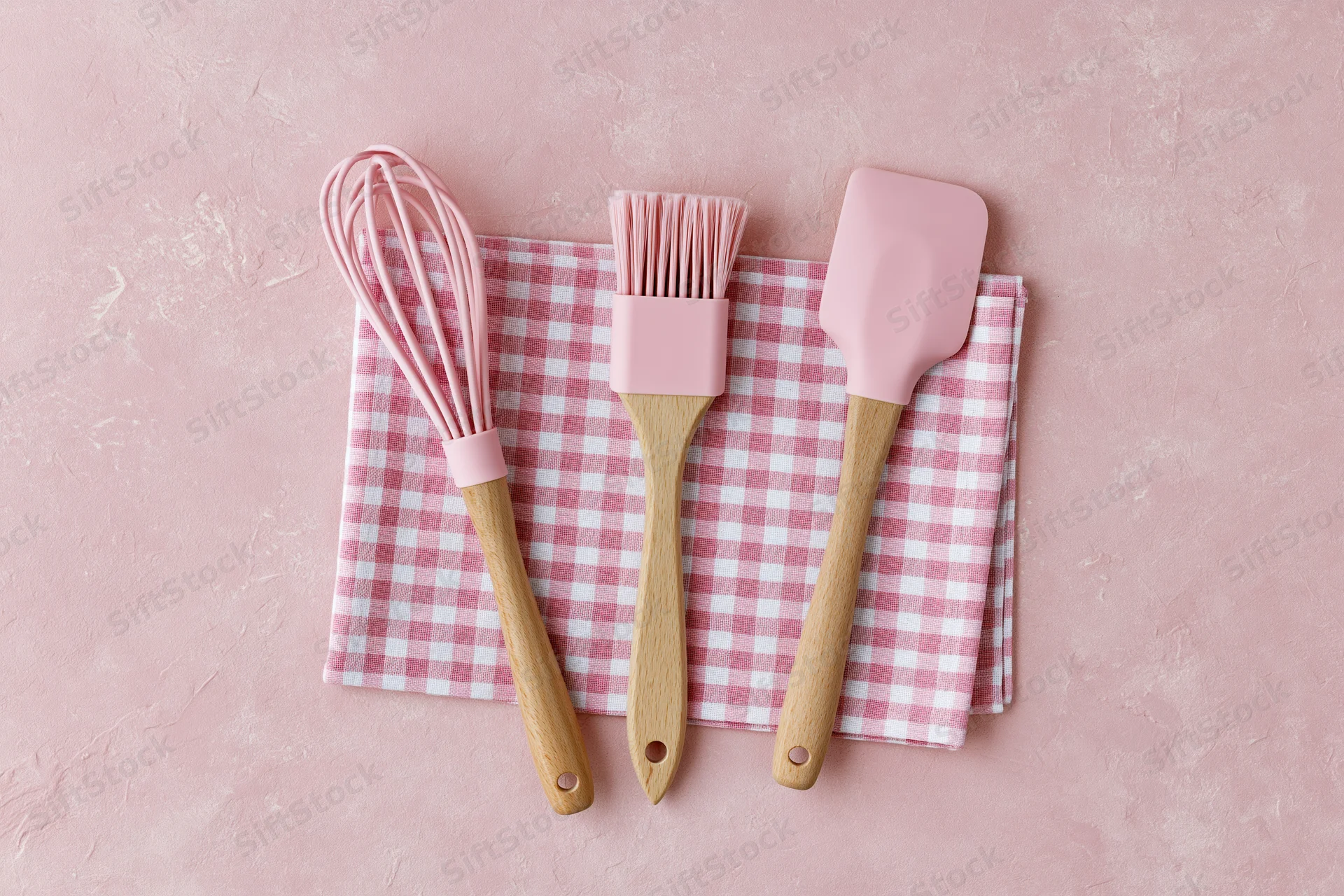 Pink silicone and wooden kitchen utensils arranged on a checkered napkin on a pink textured surface