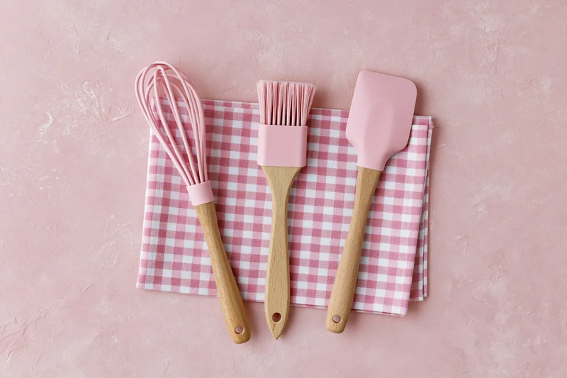 Pink silicone and wooden kitchen utensils arranged on a checkered napkin on a pink textured surface