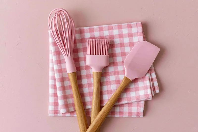 Pink silicone kitchen utensils with wooden handles arranged on a checkered napkin on a pastel pink surface