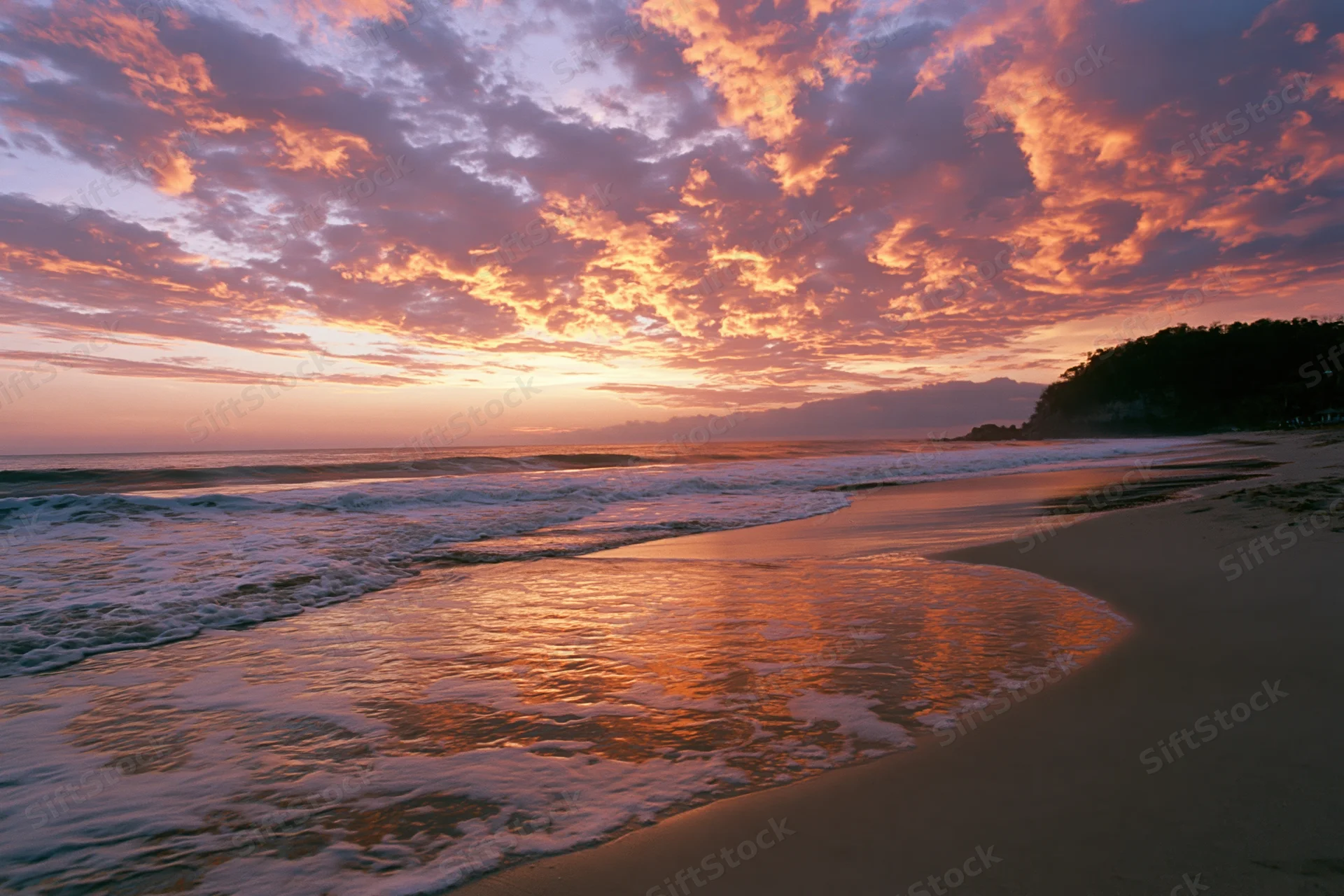 Sunset over a sandy beach with waves and colorful clouds reflecting on wet sand