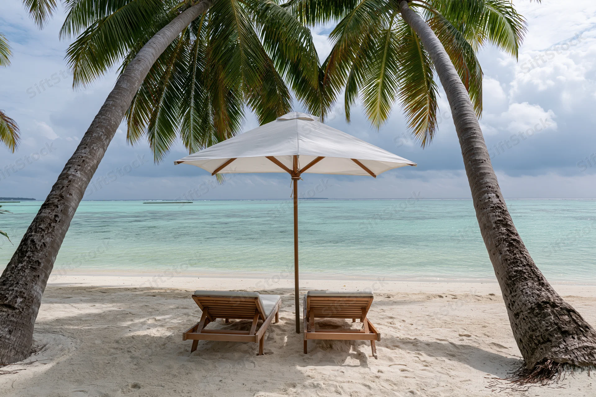 Two wooden lounge chairs under a white umbrella between palm trees on a sandy beach overlooking turquoise ocean water