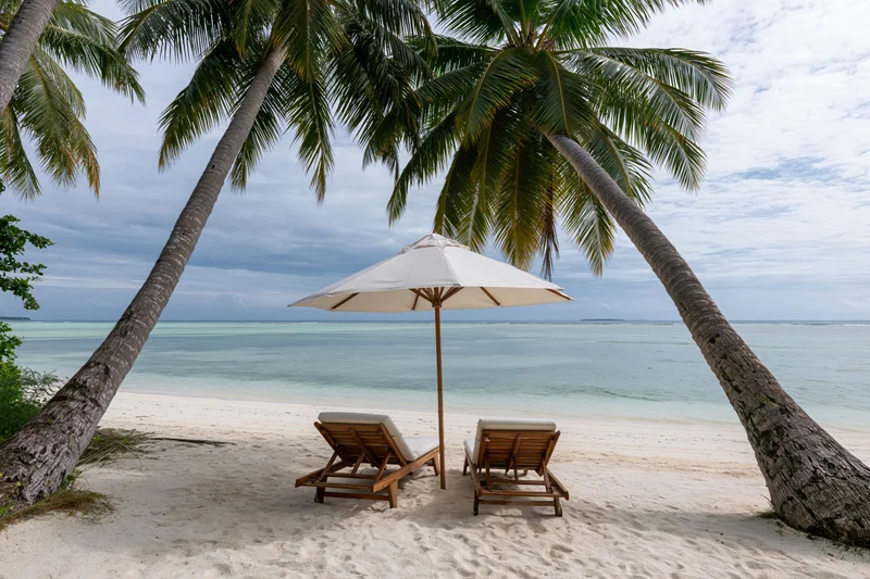 Two wooden lounge chairs under a white umbrella between palm trees on a sandy beach overlooking turquoise ocean