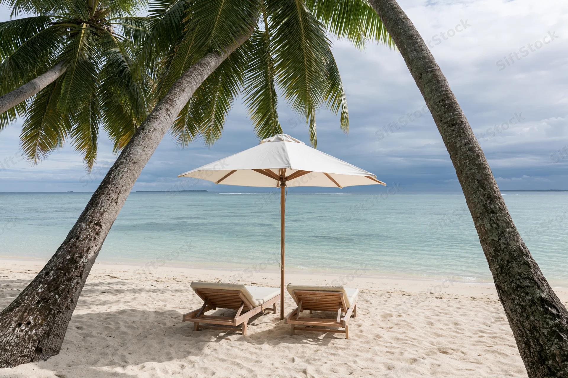 Two wooden lounge chairs under a white umbrella between palm trees on a sandy beach overlooking the ocean