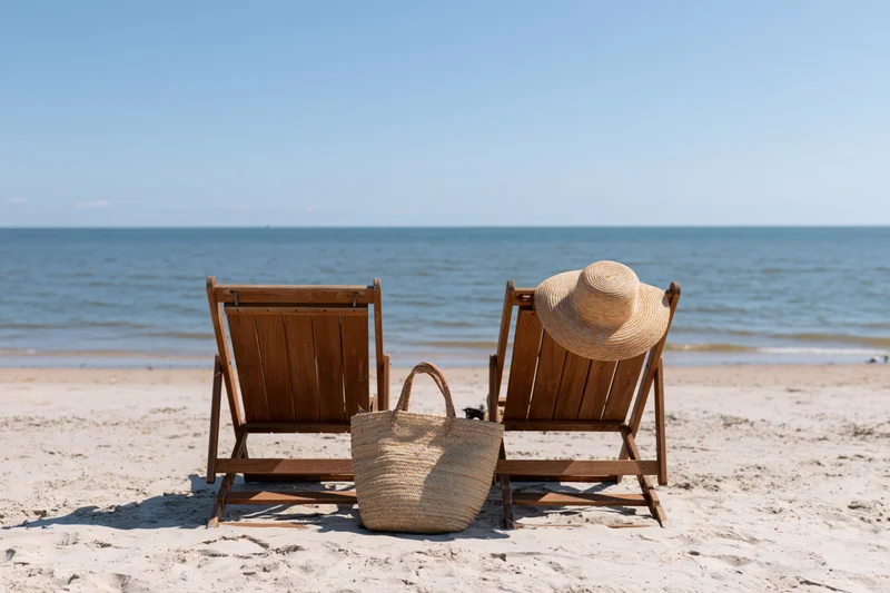 Two wooden beach chairs with a straw hat and woven bag on sandy shore facing the ocean