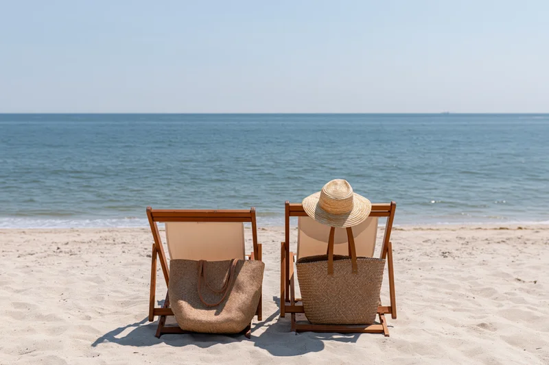 Two beach chairs with straw hat and tote bag on sandy shore facing ocean