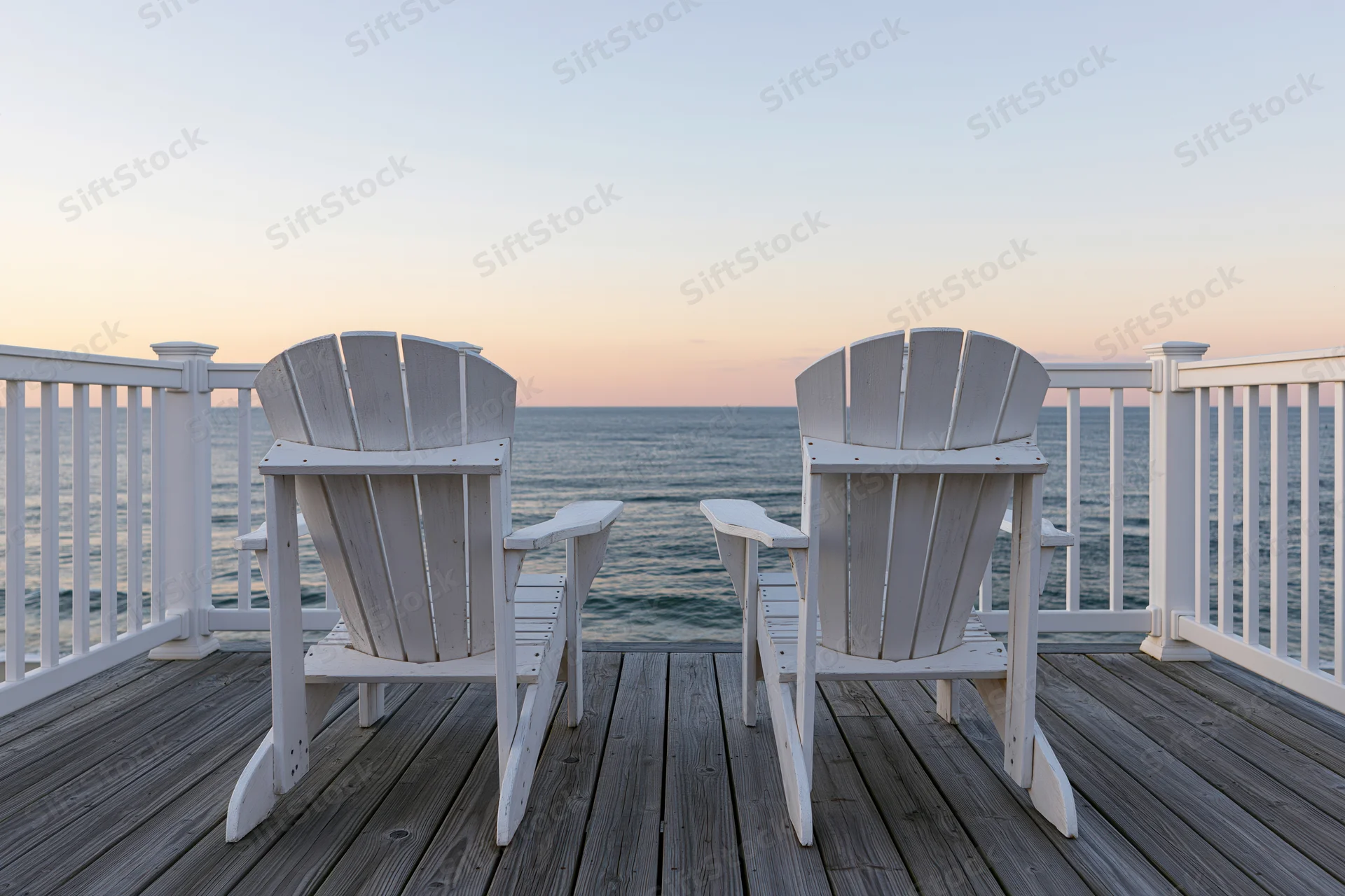 Two white adirondack chairs on a wooden deck overlooking the ocean at sunset