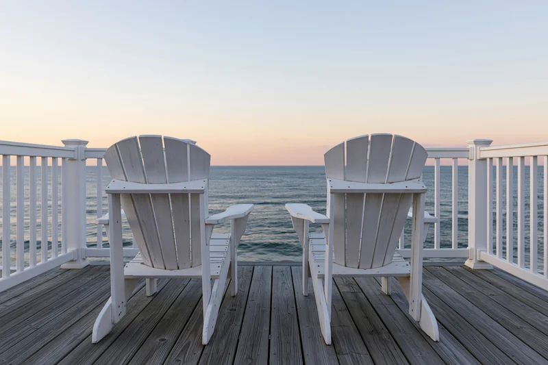Two white adirondack chairs on a wooden deck overlooking the ocean at sunset