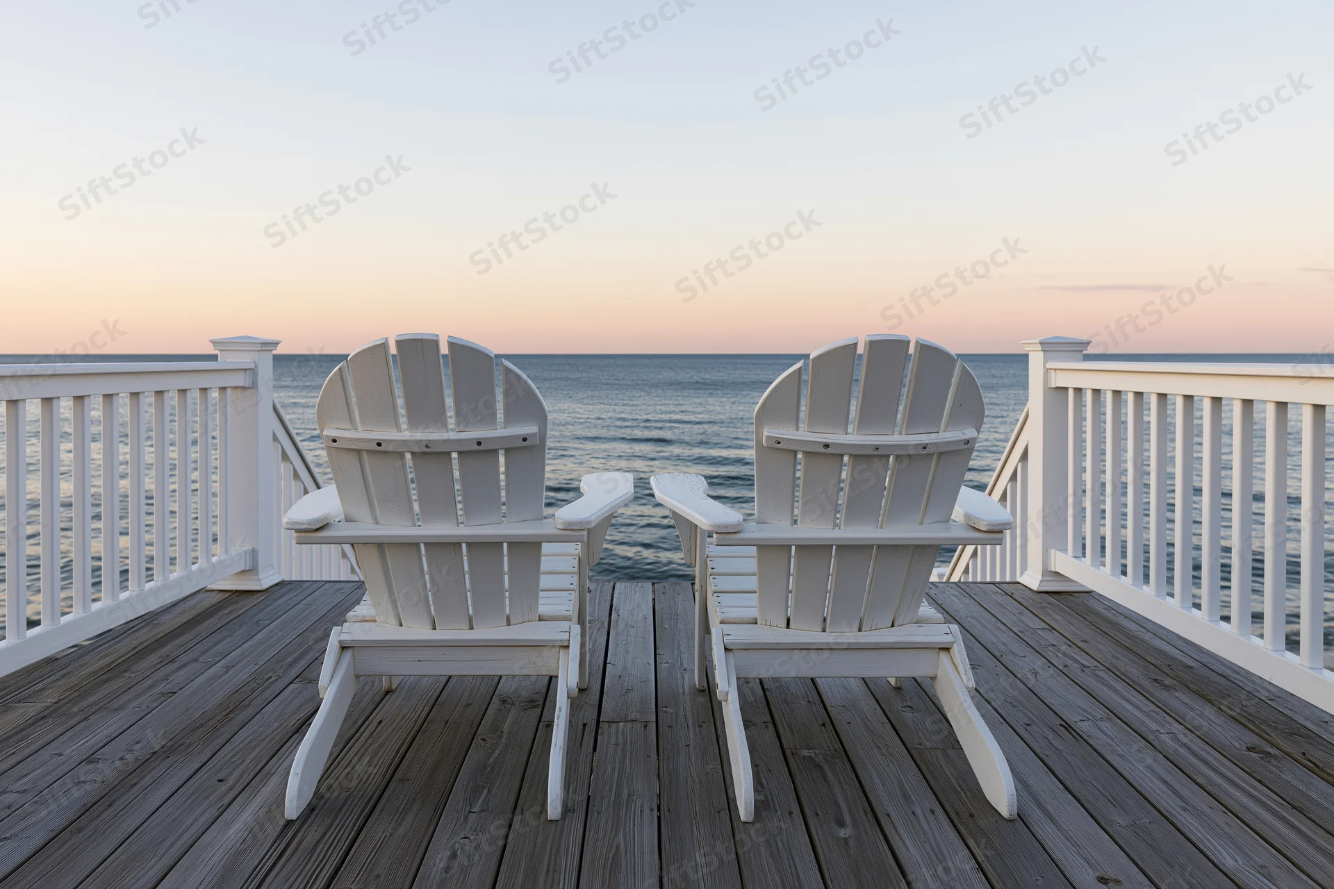 Two white adirondack chairs on a wooden deck facing the ocean at sunset