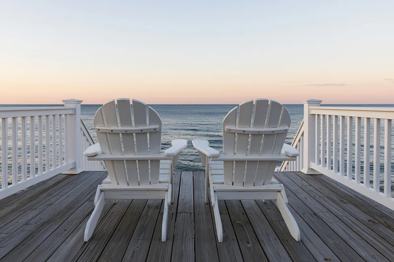 Two white adirondack chairs on a wooden deck facing the ocean at sunset