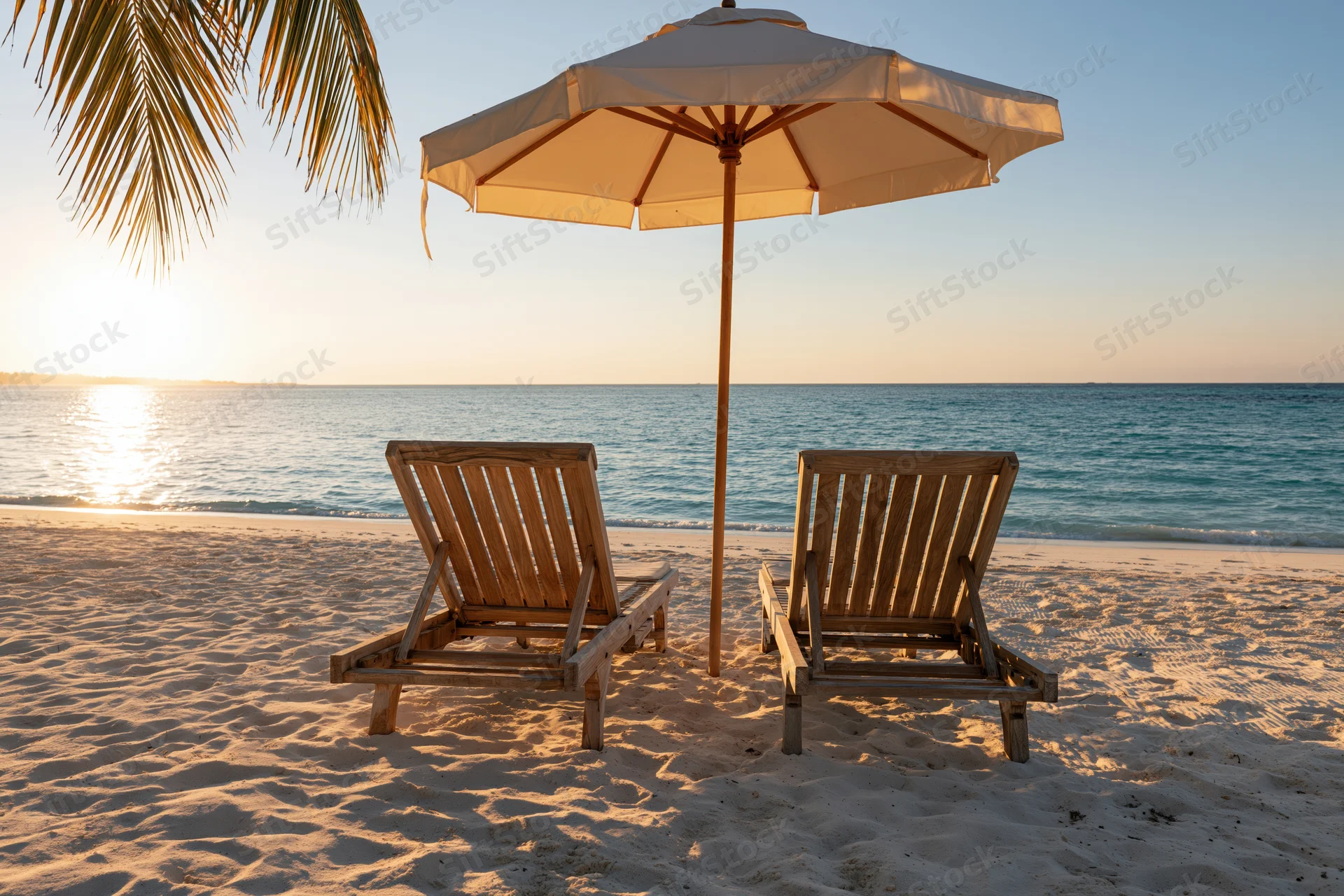 Two wooden lounge chairs under a white umbrella on a sandy beach at sunset overlooking the ocean