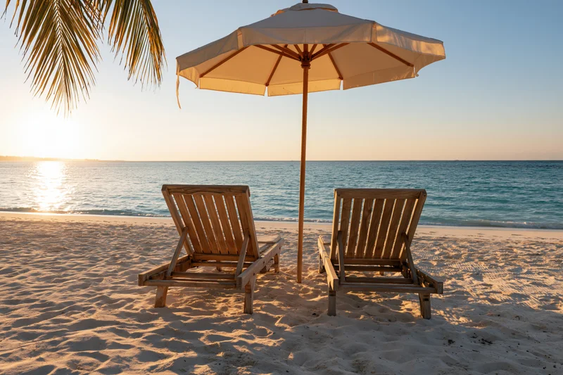 Two wooden lounge chairs under a white umbrella on a sandy beach at sunset overlooking the ocean