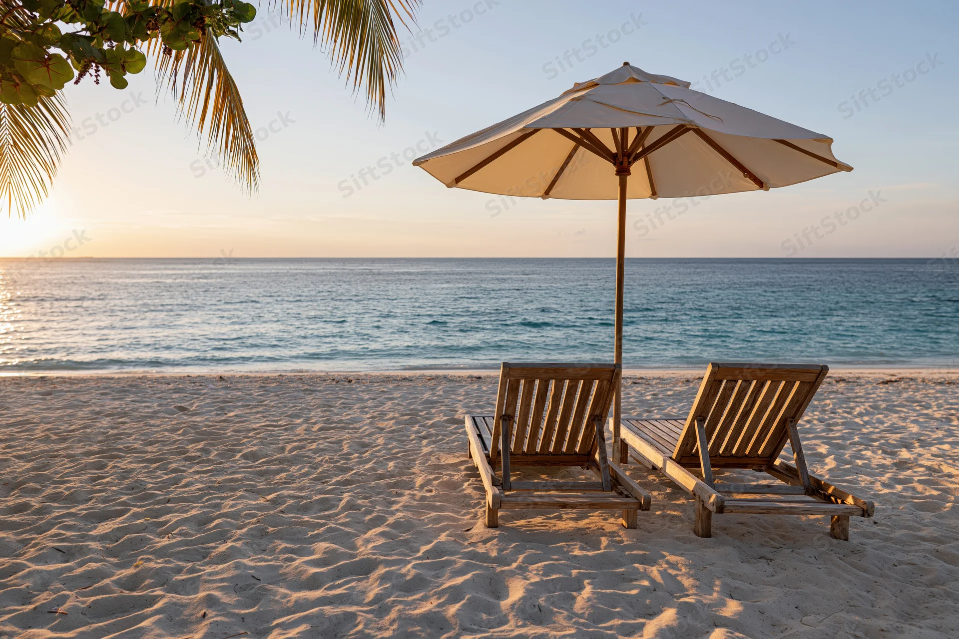 Two wooden lounge chairs under a white umbrella on a sandy beach at sunset with ocean and palm fronds in view