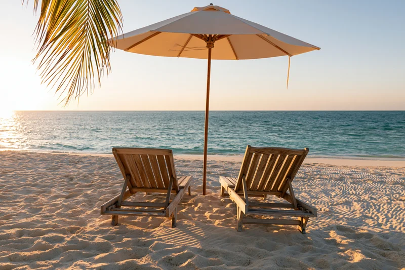 Two wooden lounge chairs under a beach umbrella on a sandy shore at sunset