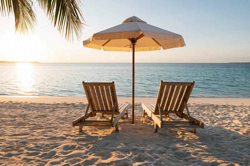 Two wooden lounge chairs under a white umbrella on a sandy beach at sunset overlooking the ocean