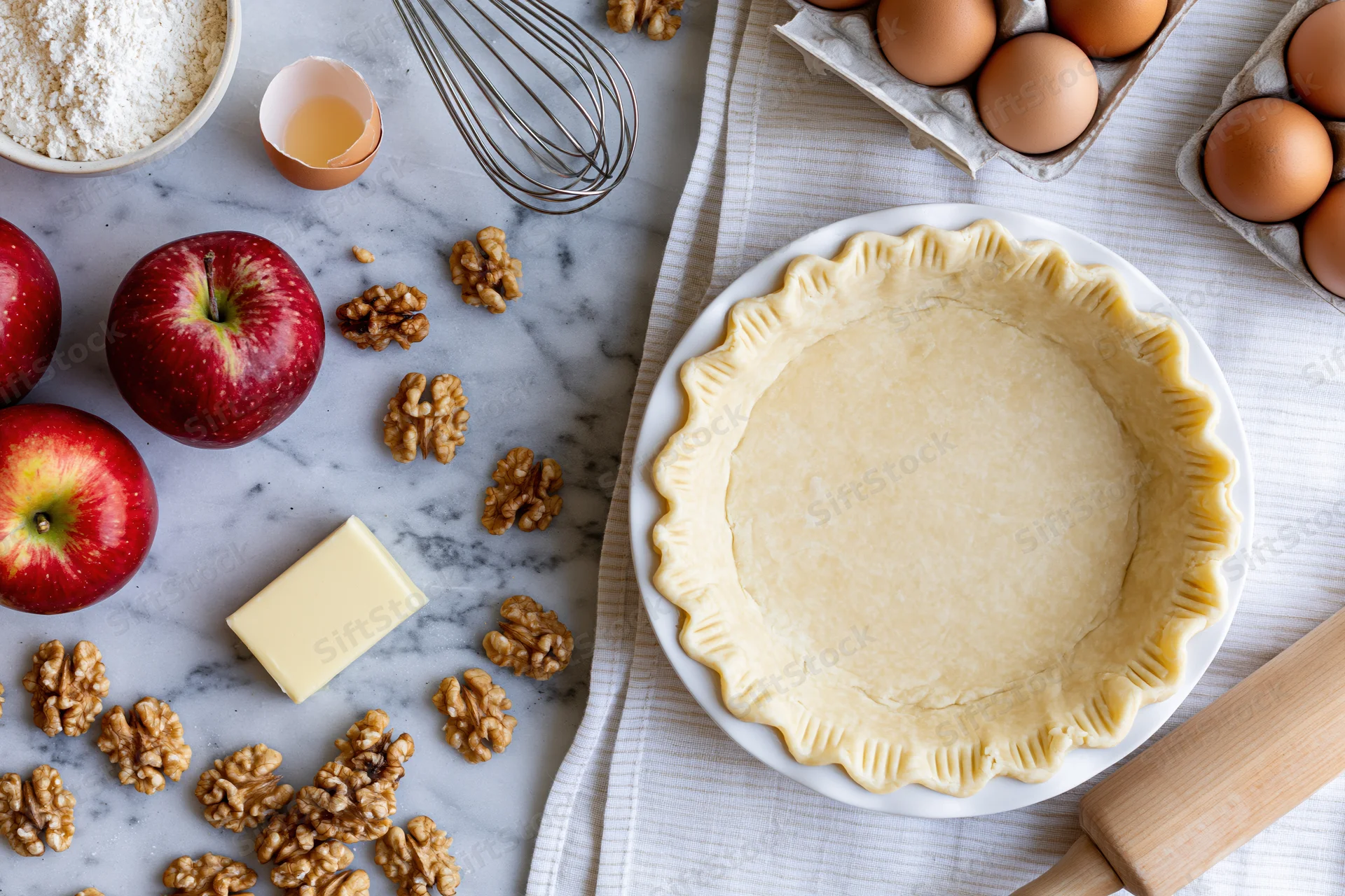 Uncooked pie crust in a white dish surrounded by apples, walnuts, eggs, flour, butter, and baking tools on a marble countertop and striped cloth