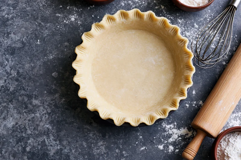 Unbaked piecrust with fluted edges on dark countertop surrounded by baking tools and flour