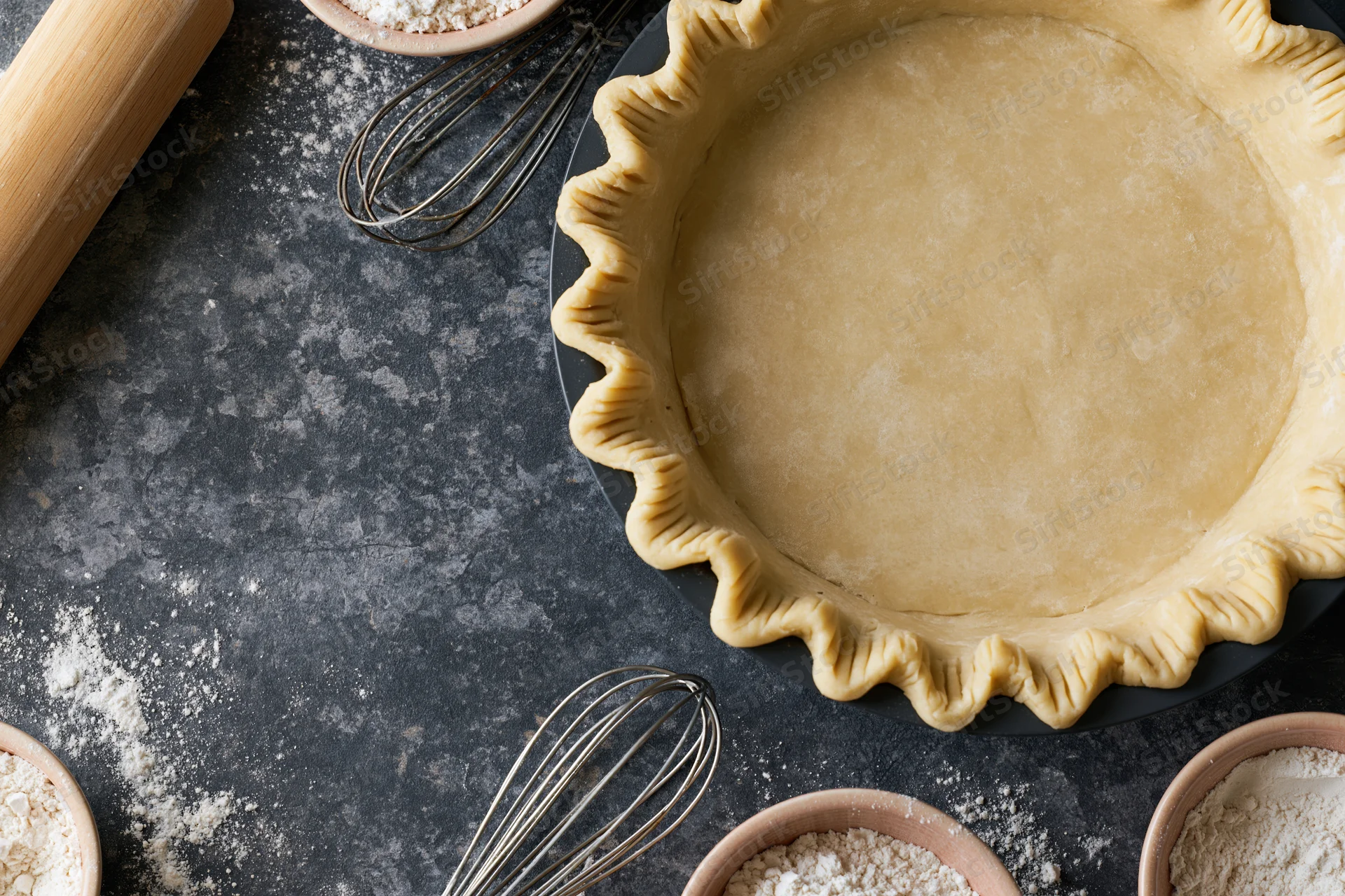 Uncooked piecrust in fluted pan on dark countertop with baking tools and flour scattered around