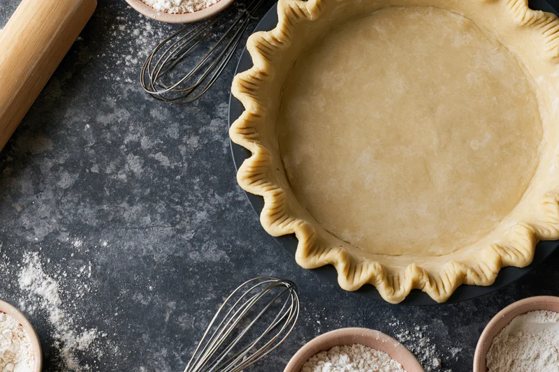 Uncooked piecrust in fluted pan on dark countertop with baking tools and flour scattered around
