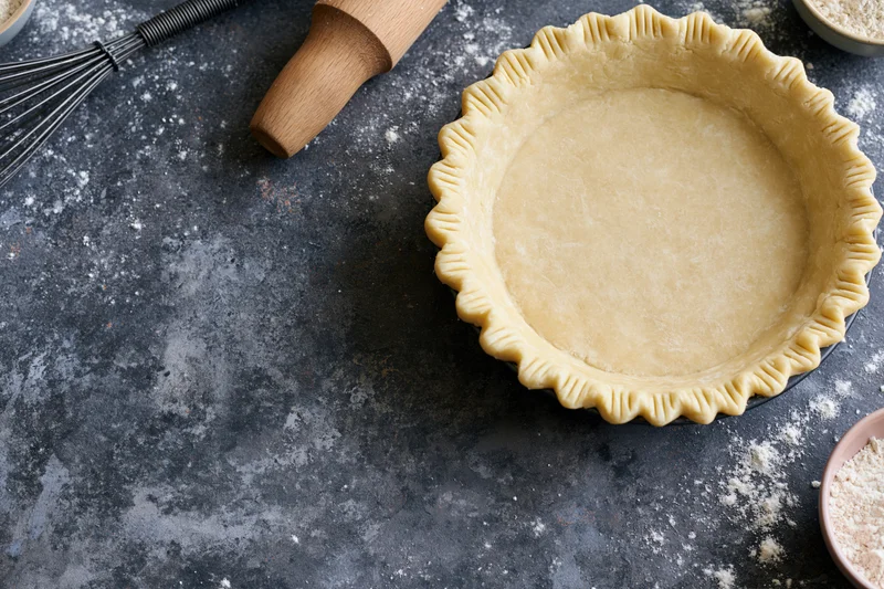 Uncooked pie crust in fluted dish on dark countertop with baking tools and flour scattered around