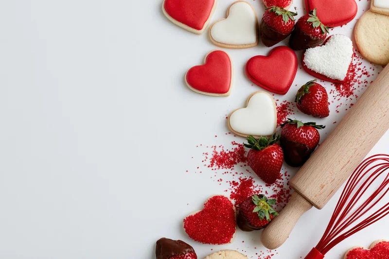 Heart-shaped cookies and strawberries with baking tools arranged on white surface