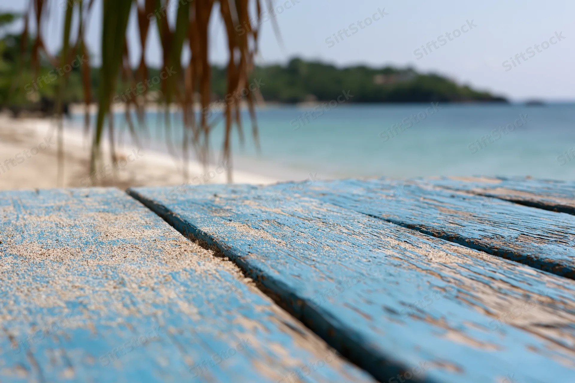Weathered blue wooden deck with sand on a tropical beach overlooking calm ocean and palm trees