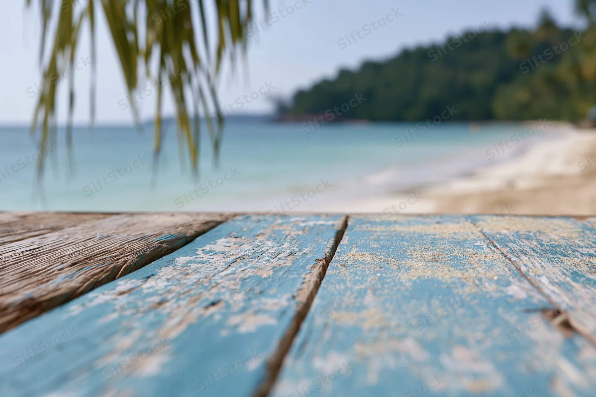 Weathered blue wooden deck with sand and palm frond overlooking turquoise ocean and tropical beach