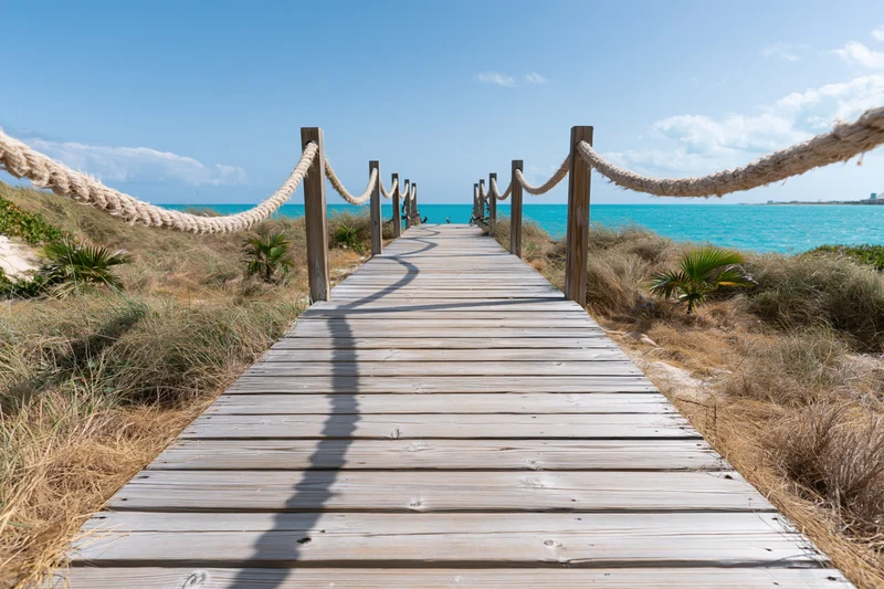 Wooden boardwalk leading to turquoise ocean along sandy dunes with rope railing
